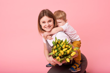 Wall Mural - Young mother hugs his little son sitting on the floor against a pink background. Mom holds a bouquet of spring yellow flowers. Care and relationships and family concept. Mothers day