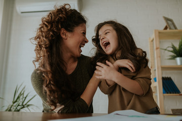 Close up of mother and daughter, beautiful brunettes, laughing at table, having fun doing homework.