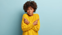 Puzzled Confused Woman Has Hesitant Expression, Curly Hairstyle, Crosses Hands Over Chest, Points Right And Left In Different Sides, Wears Yellow Sweater, Being Indifferent, Models Over Blue Wall