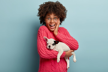 Headshot of happy black young woman gets cute pedigree dog as present from boyfriend, holds domestic little french bulldog puppy. Love and friendship between owner and animal. Heart warming photo.