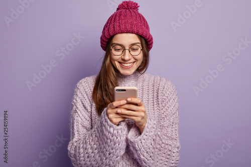 Candid shot of positive young woman plays cool game on cell phone, wastes time while waits for boyfriend, wears optical glasses, casual headgear and sweater, focused into screen, has fun indoor