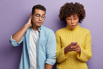 Poster - Shocked woman reads recieved notification, stares at cell phone, curious guy in round spectacles peeks at screen, wants to read message. Diverse couple with modern technolgy against purple wall