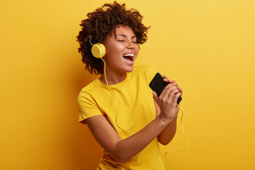 Poster - Joyful optimistic glad black woman glad to search favourite composition on web page, listens favourite cool song in headphones, sings from pleasure, has modern gadgets, wears yellow t shirt.