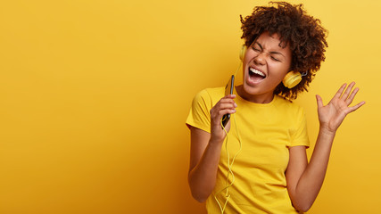 Wall Mural - Yeah, I am pleased. Joyful lovely dark skinned woman raises hand in dance move, likes great sound in new headphones, sings loudly, carries cellular, smiles broadly, wears yellow casual clothes
