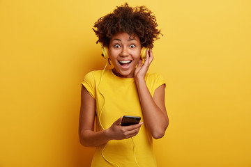 Wall Mural - Waist up shot of excited happy dark skinned young woman listens radio via cell phone and headphones, being in good mood, has break after preparing for exams, isolated over yellow background.