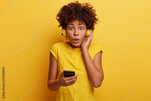 People, hobby, reaction concept. Impressed shocked dark skinned hipster girl surprised to listen audio track with high volume, has headphones on ears and smart phone in hand, isolated over yellow wall