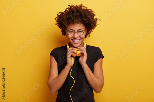 Isolated shot of optimistic woman with curly hairstyle, wears black t shirt and spectacles, feels good from spare time, enjoys listening audio book, likes hobby, being real meloman of pop music