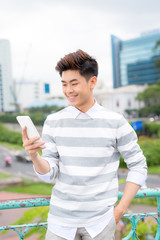 Wall Mural - Young asian handsome businessman smiling while reading his smartphone. Portrait of asian business man reading message with smart phone in outdoor.