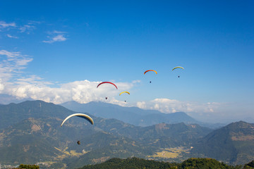 a group of paragliders on multi-colored parachutes fly over the mountain valley in a clear blue sky with white clouds