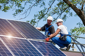 two young technicians mounting heavy solar photo voltaic panel on tall steel platform on green tree 