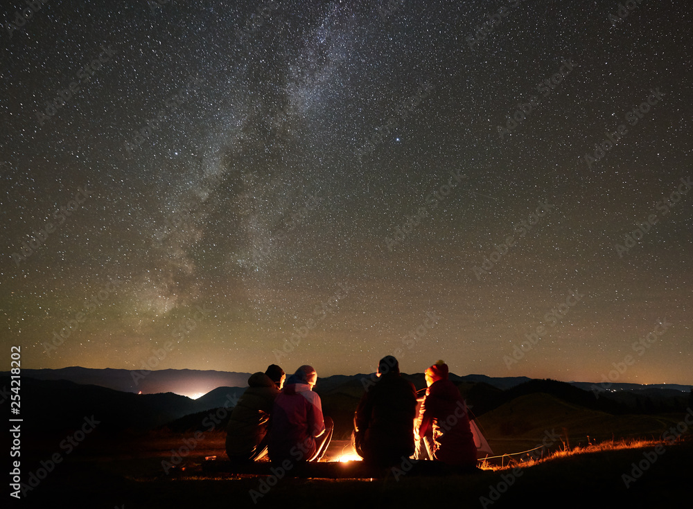 Night summer camping in the mountains. Back view group of four friends tourists sitting on a bench made of logs together around campfire under amazing night starry sky full of stars and Milky way. - obrazy, fototapety, plakaty 