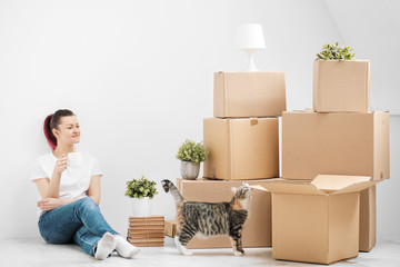 Wall Mural - A young beautiful brunette girl in a white T-shirt is sitting on the floor of a bright room and making calls on her smartphone. Around cardboard boxes and a cat.