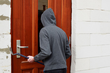 Robber in hood trying open door with crowbar and looking through glass pane, back view. Crime, burglary and house robbery concept.