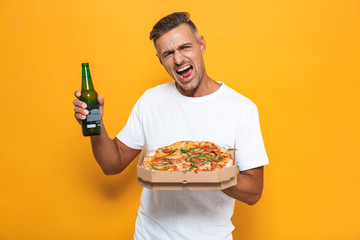 Canvas Print - Image of excited man 30s in white t-shirt drinking beer and eating pizza while standing isolated