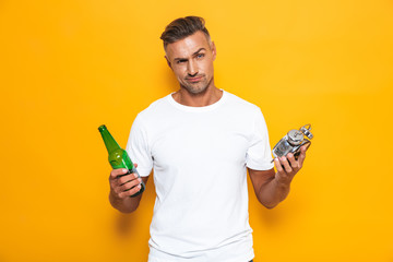 Poster - Image of bearded man 30s in white t-shirt drinking beer and holding alarm clock while standing isolated