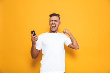 Wall Mural - Image of attractive man 30s in white t-shirt smiling and holding smartphone while standing isolated