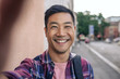 © mavoimages - Smiling young Asian man taking selfies on a city street