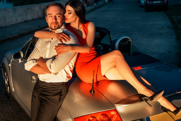 Gorgeous sexy copule of man and woman on a vacation car trip in summer day. Luxury grey sport car. Sexy fit brunette woman and man in evening outfit. Night portrait