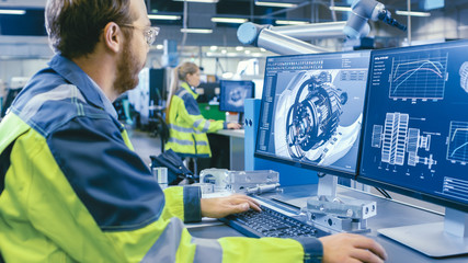 Wall Mural - At the Factory: Mechanical Engineer Works on Computer, Designs in CAD 3D Model of the Engine. In the Background Factory Workers and Manufacturing Process.