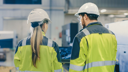 Fototapete - Male Industrial Worker and Female Chief Mechanical Engineer in Walk Through Manufacturing Plant while Discuss Factory's New Project and Using Laptop. Facility Has Working Machinery.