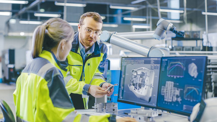 Fototapete - At the Factory: Male Mechanical Engineer Holds Component and Female Chief Engineer Work on Personal Computer, They Discuss Details of the 3D Engine Model Design for Robotic Arm.