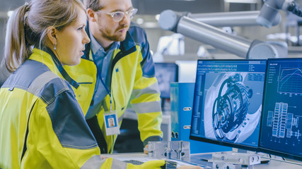 Wall Mural - At the Factory: Male Mechanical Engineer and Female Chief Engineer Work Together on the Personal Computer, They Discuss Details of the 3D Engine Model for Robotic Arm.