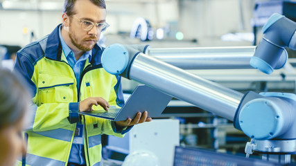 Fototapete - At the Factory: Automation Engineer Uses Laptop for Programming Robotic Arm. New Era in Automatic Manufacturing Industry.