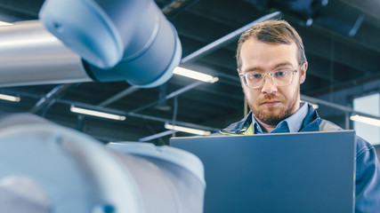 Fototapete - Low Angle Shot At the Factory: Automation Engineer Uses Laptop for Programming and Testing Robotic Arm. New Era in Automatic Manufacturing Industry.