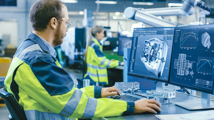 Fototapete - At the Factory: Mechanical Engineer Works on Computer, Designs in CAD 3D Model of the Engine. In the Background Factory Workers and Manufacturing Process.