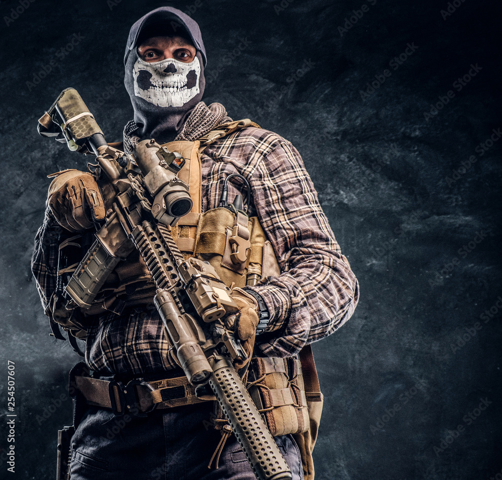 Obraz Private security service contractor wearing a balaclava skull and cap holding an assault. Studio photo against a dark textured wall fototapeta, plakat