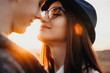 © Strelciuc - Close up portrait of a beautiful woman with dark hair wearing hat and glasses smiling and trying to kiss her boyfriend against sunset while dating outside.