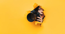 A Paparazzi Girl With A Camera Looks Out From Cover And Looks At What Is Happening With Her Mouth Open. Yellow Paper, Torn Hole. Tabloid Press. Copy Space.