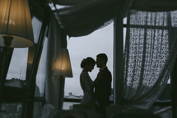 beautiful and elegant blonde bride in a long white dress with her handsome bride in a blue suit standing in the hotel room near window