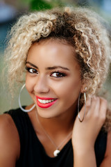 Wall Mural - Emotional cheerful beautiful afro american woman with curly hair enjoying leisure time while looking at camera sitting ain park .