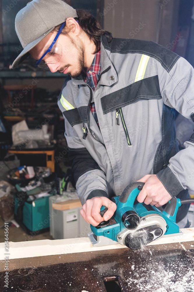 A young carpenter with a beard works with an electric plane without suctioning sawdust. Leveling and sanding wooden beams
