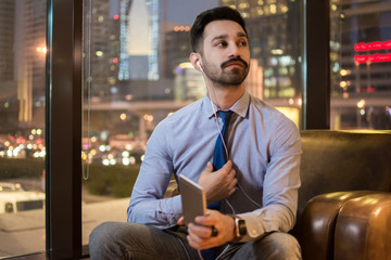 Wall Mural - Handsome young businessman with earphones listening to music on smartphone while sitting in comfortable sofa with urban buildings in the background at night