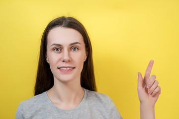 Closeup portrait of beautiful cheerful young girl with smile, being in great mood, happy to show something upwards, poses against yellow background, dressed in gray T-shirt