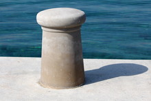 New Dark Grey Concrete Mooring Bollard On Top Of Stone And Concrete Pier With Calm Clear Blue Sea In Background On Warm Spring Day