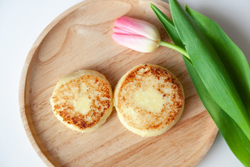 Closeup fresh pink tulip on wooden round plate, two golden bitten fried cheesecakes in the shape of figure 8, dessert made from cottage cheese. Concept tasty and healthy breakfast March 8