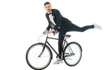 Wall Mural - cheerful groom in elegant suit making stunts on bike isolated on white