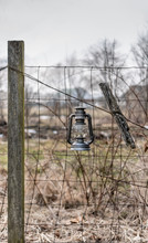 Vintage Oil Lamp Lantern Hanging On Farmers Wire Fence Post With Field In Background