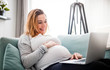 © leszekglasner - Smiling pregnant woman using laptop, sitting on sofa at home