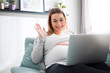 © leszekglasner - Pregnant woman video chatting with family on laptop waving hand to screen