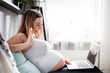 © leszekglasner - Pregnant woman video chatting with family on laptop waving hand to screen