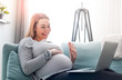 © leszekglasner - Pregnant woman video chatting with family on laptop waving hand to screen