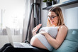 © leszekglasner - Pregnant woman video chatting with family on laptop waving hand to screen