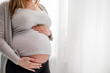 © leszekglasner - Pregnant woman touching her belly standing by the window at home