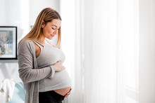 Beautiful Pregnant Woman Touching Her Belly Standing By The Window At Home