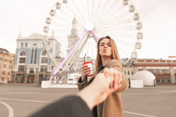 Male hand holds a stylish girl, she kisses with a coffee in her hand. Girl holds her boyfriend's hand and makes an air kiss against the background of the city landscape. Love. Follow me