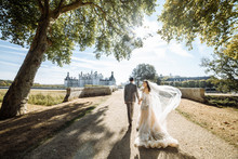 Beautiful Photo Of Happy Newlywed Couple. Bride In A Luxurious Dress With A Long Bridal Veil. They Go To The Old Beautiful Castle In France